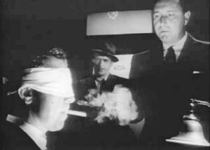 Philip Marlowe with his eyes bandaged being interrogated by Police Lieutenant Randall (Don Douglas) about two murders.